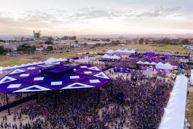 Vista aérea de festival ao ar livre ao entardecer com grande público, palco com cobertura roxa em formato geométrico, tendas brancas e pessoas vestindo camisetas roxas