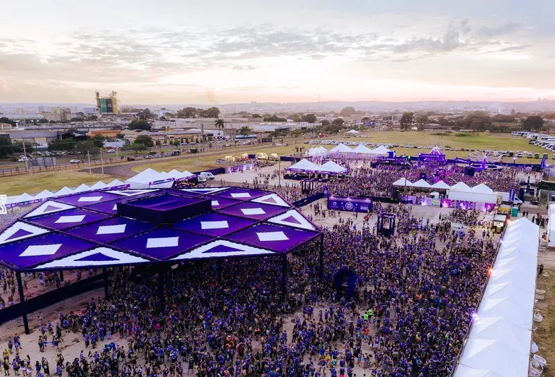 Vista aérea de festival ao ar livre ao entardecer com grande público, palco com cobertura roxa em formato geométrico, tendas brancas e pessoas vestindo camisetas roxas