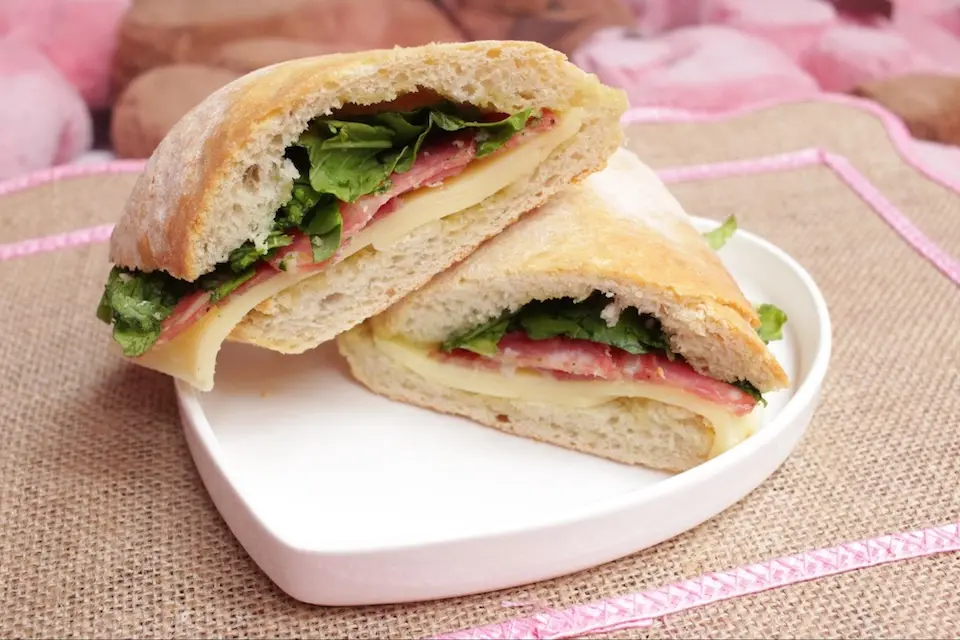 ciabatta cortada ao meio com fatias de salame, queijo e rúcula sobre prato branco