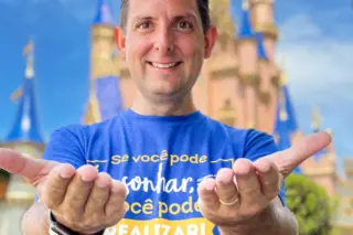 Homem adulto sorrindo, de frente, com as mãos estendidas e camiseta azul com texto, castelo com torres ao fundo