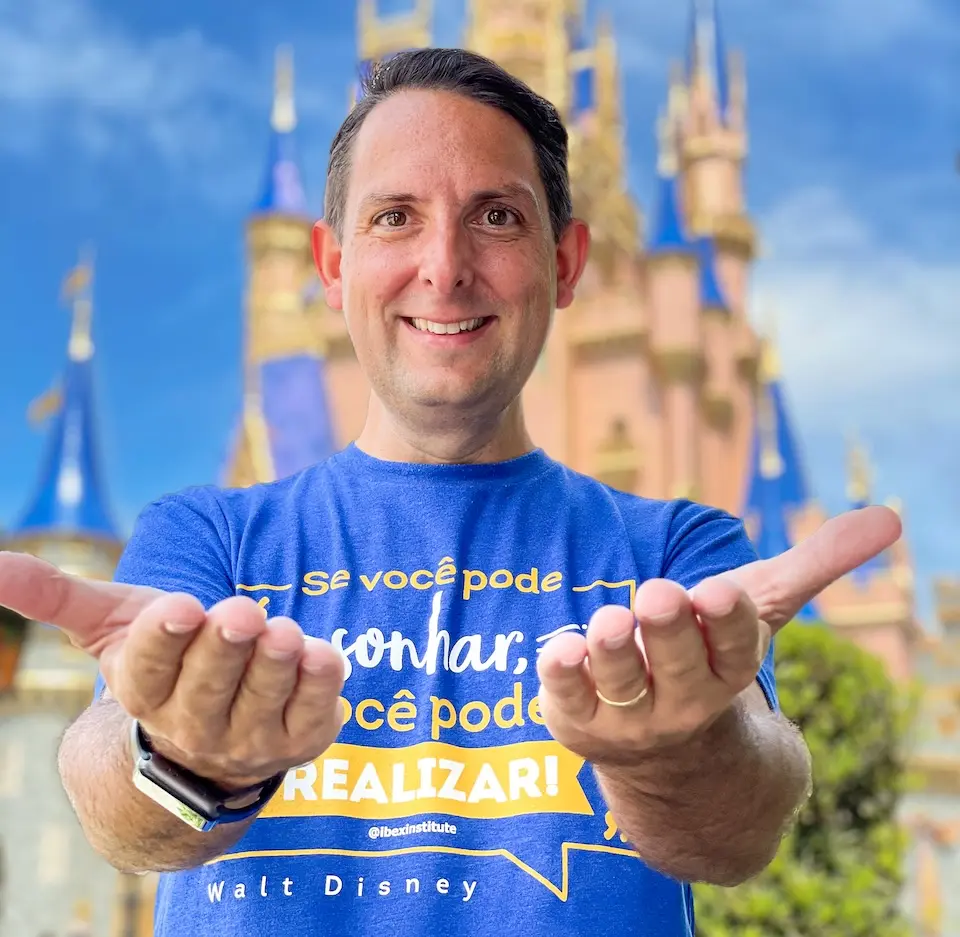 Homem adulto sorrindo, de frente, com as mãos estendidas e camiseta azul com texto, castelo com torres ao fundo