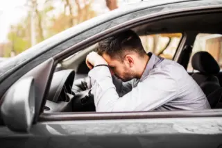 homem sentado no banco do motorista com a cabeça apoiada nos braços sobre o volante