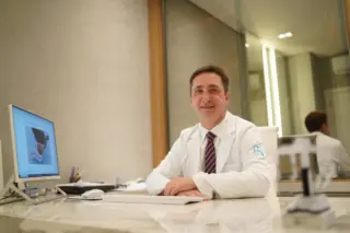 Homem adulto de jaleco branco sentado atrás de mesa de consultório, com monitor de computador à esquerda e reflexo no vidro ao fundo