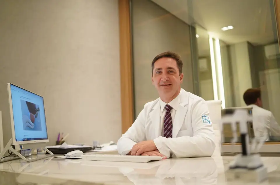 Homem adulto de jaleco branco sentado atrás de mesa de consultório, com monitor de computador à esquerda e reflexo no vidro ao fundo