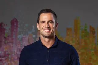 Homem sorrindo com camisa azul escura, retrato em meio corpo frente a fundo colorido com formas que lembram prédios
