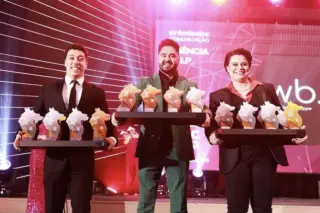 Três profissionais exibem troféus durante cerimônia de premiação do setor de comunicação.