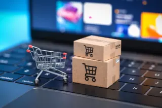 Mini carrinho de compras metálico e duas caixas de papelão com ícones de carrinho sobre o teclado de um notebook, com tela desfocada ao fundo