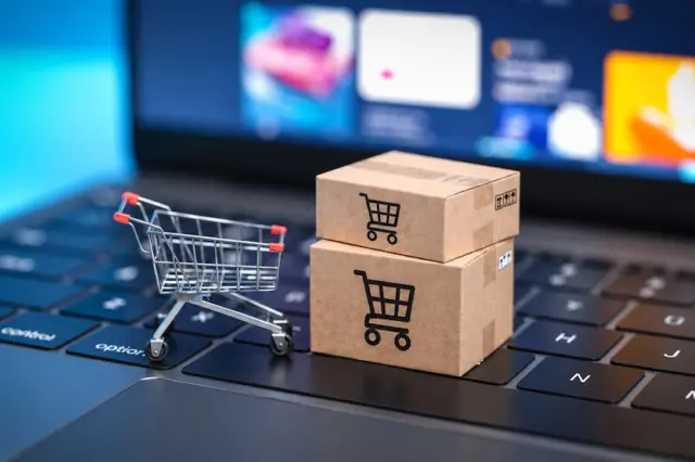 Mini carrinho de compras metálico e duas caixas de papelão com ícones de carrinho sobre o teclado de um notebook, com tela desfocada ao fundo