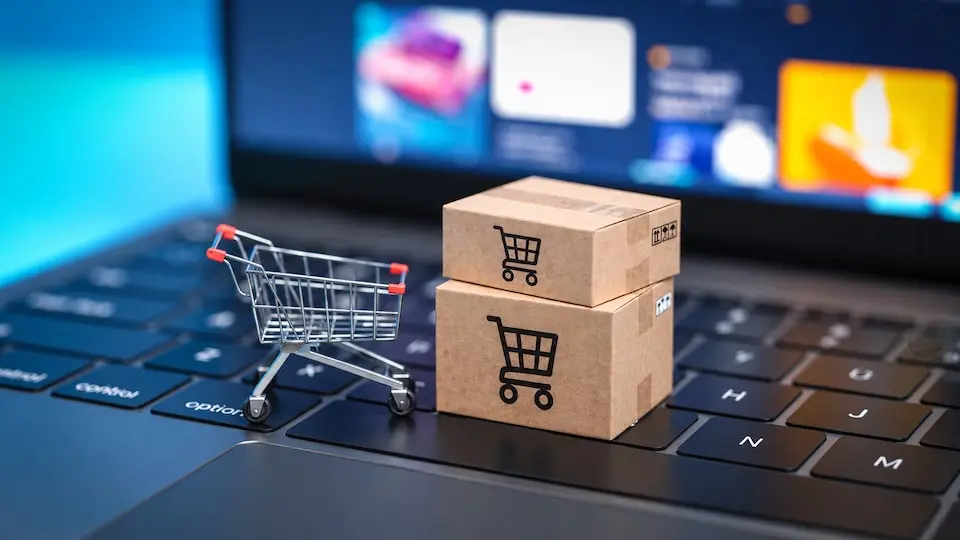Mini carrinho de compras metálico e duas caixas de papelão com ícones de carrinho sobre o teclado de um notebook, com tela desfocada ao fundo