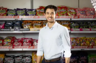 homem adulto sorrindo em pé diante de prateleiras cheias de embalagens de doces coloridas