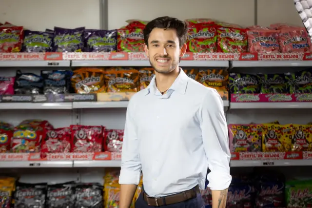 homem adulto sorrindo em pé diante de prateleiras cheias de embalagens de doces coloridas