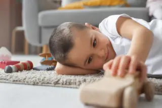 criança deitada de lado sobre tapete claro brincando com um carrinho de madeira, com sofá desfocado ao fundo