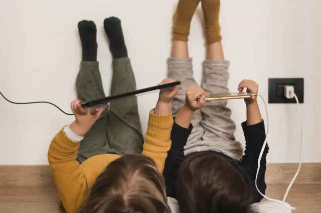 duas crianças deitadas no chão com as pernas apoiadas na parede, segurando um tablet e um celular conectado a uma tomada