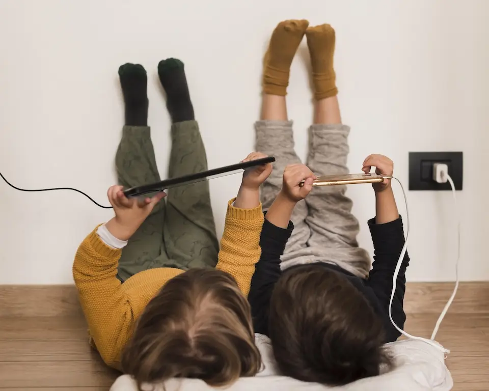 duas crianças deitadas no chão com as pernas apoiadas na parede, segurando um tablet e um celular conectado a uma tomada