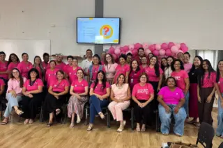 Grupo de participantes em sala, a maioria vestindo camisetas rosas, com painel de balões cor-de-rosa e uma tela de apresentação ao fundo