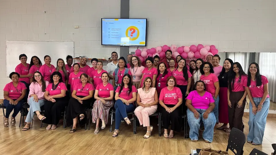 Grupo de participantes em sala, a maioria vestindo camisetas rosas, com painel de balões cor-de-rosa e uma tela de apresentação ao fundo