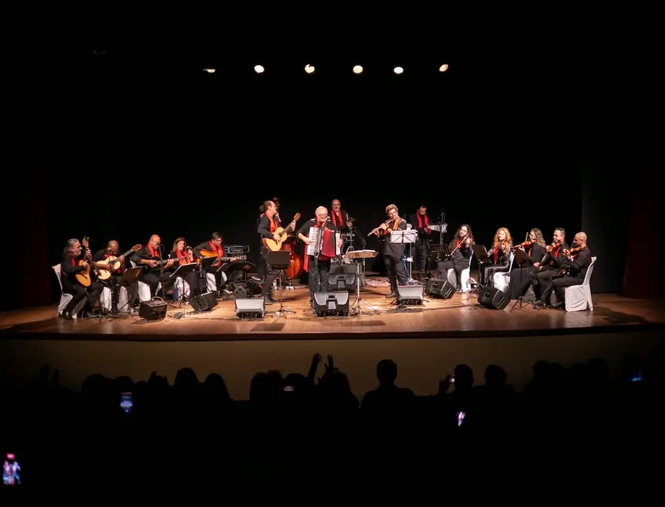 Visão ampla do palco durante apresentação com músicos sentados e em pé, instrumentos de cordas e público em silhueta
