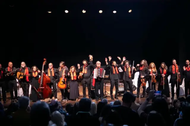Grupo de músicos alinhados no palco segurando diversos instrumentos de cordas e um acordeão, com lenços ou faixas laranja no pescoço