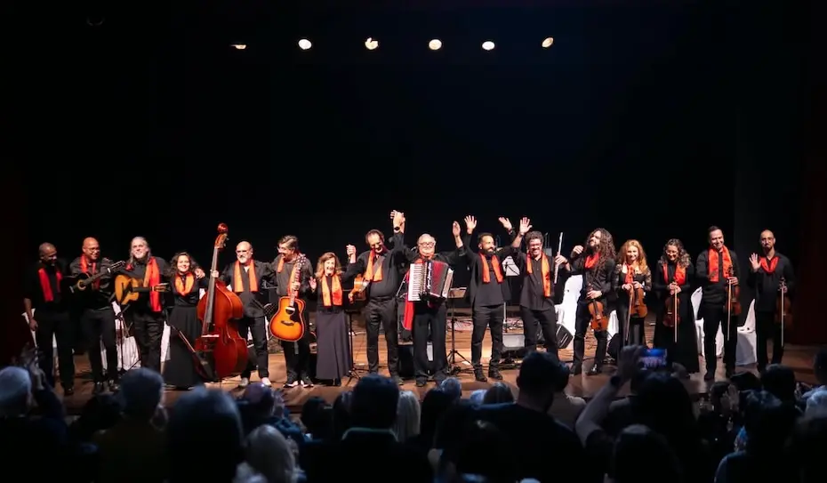 Grupo de músicos alinhados no palco segurando diversos instrumentos de cordas e um acordeão, com lenços ou faixas laranja no pescoço