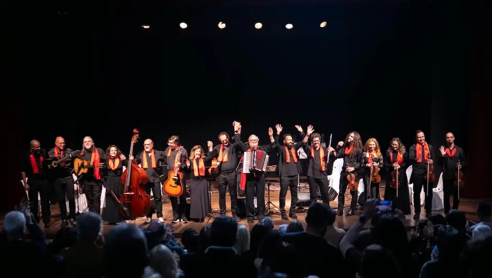 Grupo de músicos alinhados no palco segurando diversos instrumentos de cordas e um acordeão, com lenços ou faixas laranja no pescoço