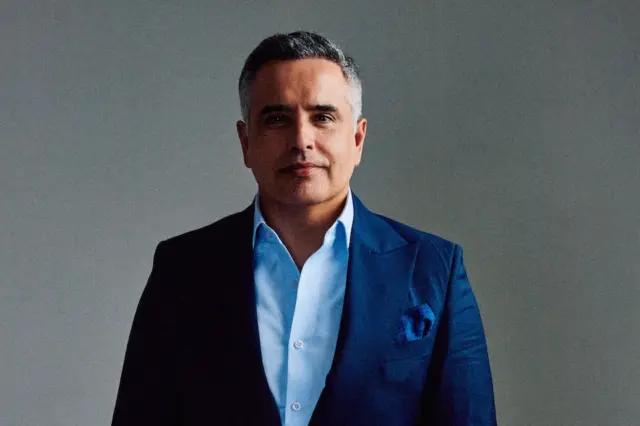 Homem adulto em terno azul e camisa azul-clara, olhando para a câmera, em retrato com fundo cinza