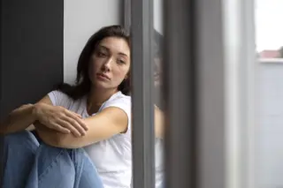 mulher sentada ao lado de uma janela, olhando pensativa com reflexo no vidro
