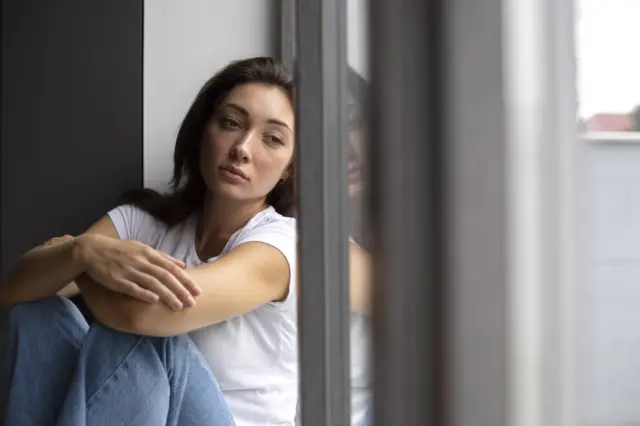 mulher sentada ao lado de uma janela, olhando pensativa com reflexo no vidro