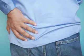 Pessoa segurando a região lombar com a mão direita, usando camisa azul clara e calça jeans, fundo azul-esverdeado