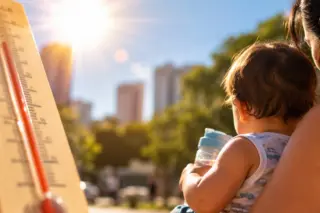 Termômetro com coluna vermelha em primeiro plano, sol forte ao fundo; adulto de costas segurando bebê com mamadeira e prédios desfocados ao fundo