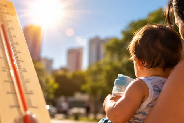 Termômetro com coluna vermelha em primeiro plano, sol forte ao fundo; adulto de costas segurando bebê com mamadeira e prédios desfocados ao fundo