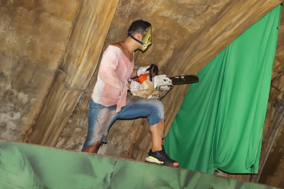 Pessoa fantasiada com máscara dourada segurando uma motosserra sobre plataforma, com cortina verde ao fundo