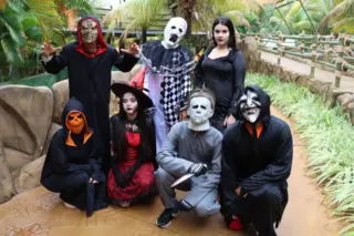 Sete pessoas fantasiadas com máscaras e roupas de Halloween posam em um percurso do parque aquático com vegetação e pedras artificiais ao fundo