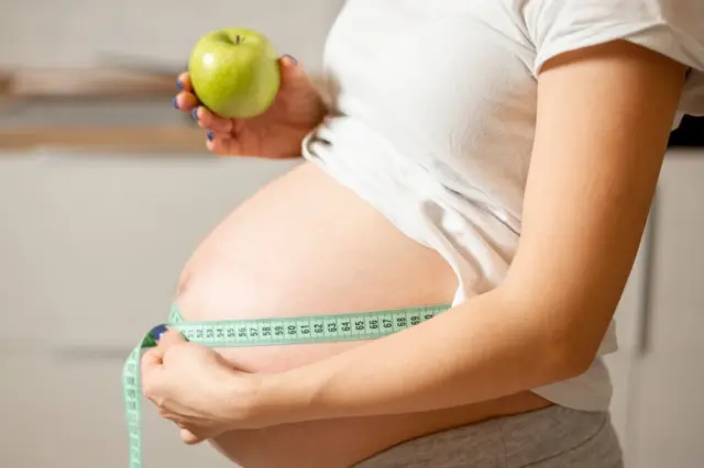 barriga de mulher grávida sendo medida com fita métrica verde; mão segura uma maçã verde