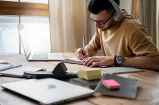 Pessoa com óculos e fones de ouvido escrevendo em caderno ao lado de laptop e blocos de notas sobre mesa de madeira