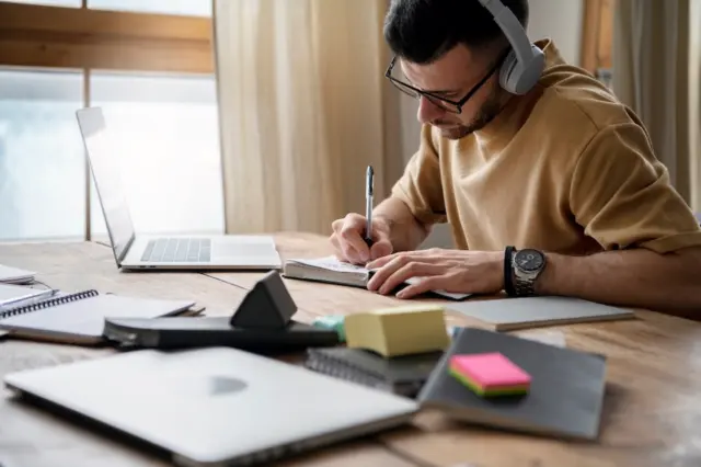 Pessoa com óculos e fones de ouvido escrevendo em caderno ao lado de laptop e blocos de notas sobre mesa de madeira