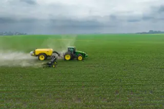 Trator verde puxando um tanque amarelo aplicando insumo sobre uma vasta lavoura de mudas sob céu nublado