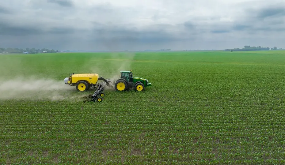 Trator verde puxando um tanque amarelo aplicando insumo sobre uma vasta lavoura de mudas sob céu nublado