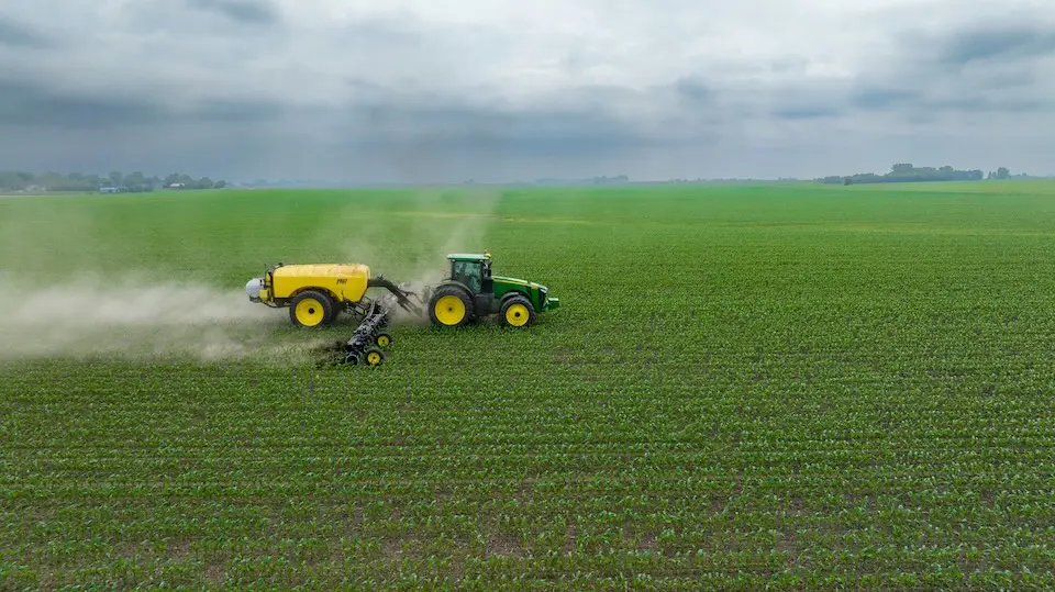 Trator verde puxando um tanque amarelo aplicando insumo sobre uma vasta lavoura de mudas sob céu nublado