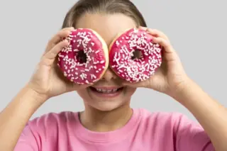 criança sorrindo segura dois donuts com cobertura rosa e granulados cobrindo os olhos