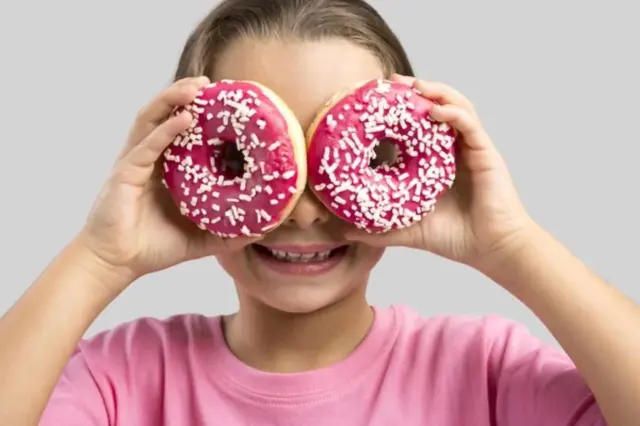 criança sorrindo segura dois donuts com cobertura rosa e granulados cobrindo os olhos
