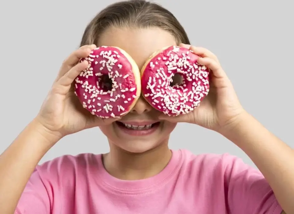 criança sorrindo segura dois donuts com cobertura rosa e granulados cobrindo os olhos