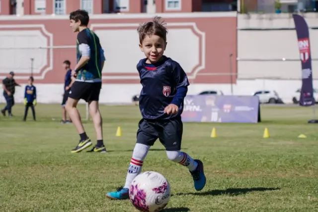 criança correndo em campo gramado enquanto avança em direção a uma bola branca com detalhes rosa; outras pessoas e um prédio ao fundo