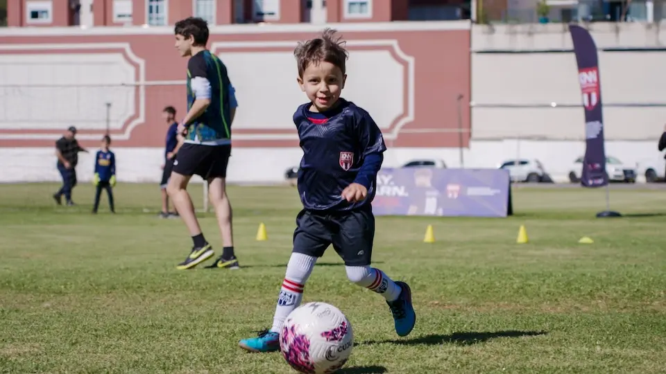 criança correndo em campo gramado enquanto avança em direção a uma bola branca com detalhes rosa; outras pessoas e um prédio ao fundo