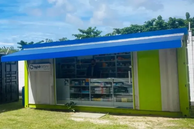 Quiosque verde com toldo azul e vitrine de farmácia com prateleiras de produtos, em área gramada