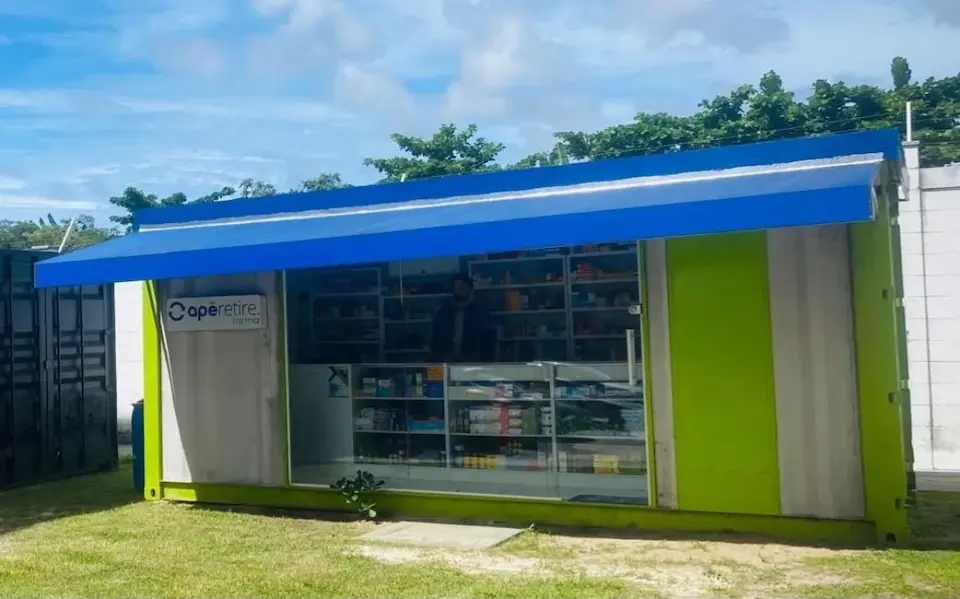 Quiosque verde com toldo azul e vitrine de farmácia com prateleiras de produtos, em área gramada