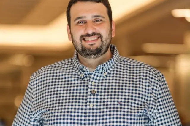 homem sorridente com barba curta e camisa xadrez azul e branca em ambiente interno com fundo desfocado