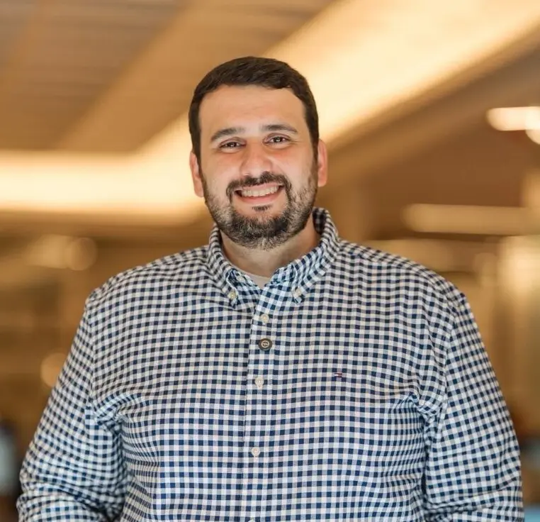 homem sorridente com barba curta e camisa xadrez azul e branca em ambiente interno com fundo desfocado