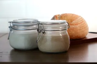 Dois potes de vidro com massa madre e um pão redondo rústico ao fundo sobre bancada de madeira