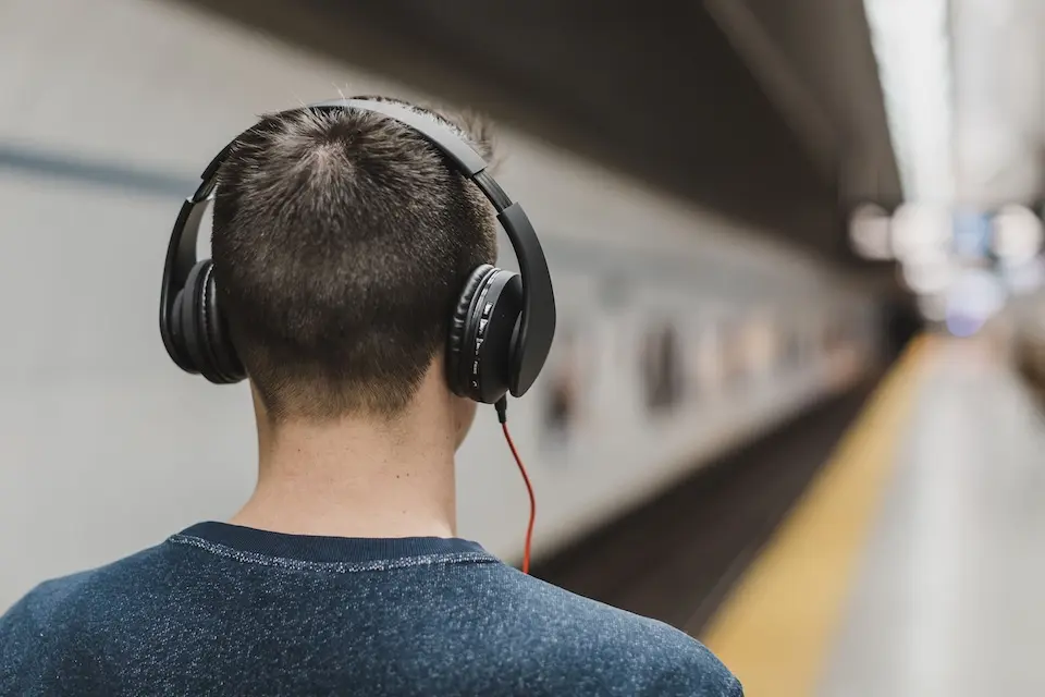Pessoa vista por trás usando fone de ouvido circumaural preto com fio vermelho numa plataforma de estação com trilhos desfocados ao fundo