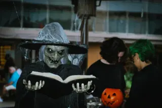 bruxa fantasiada com rosto esverdeado segurando um livro aberto, com duas pessoas ao fundo e um balde laranja com rosto de abóbora